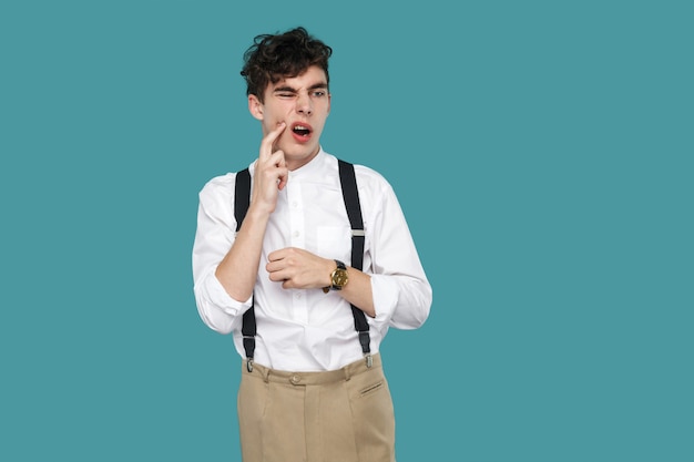 Dor de dente. homem tocando seu queixo por causa da dor de dente. retrato de jovem empresário encaracolado hipster bonito no clássico casual camisa branca e suspensório em pé. estúdio tiro isolado sobre fundo azul.