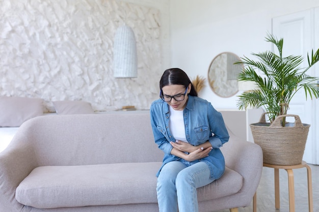 Dor de estômago jovem linda de óculos e jeans sentado no sofá em casa e segurando-a