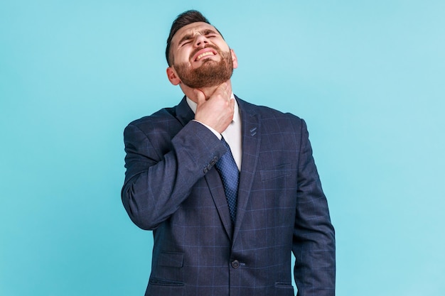 Dor de garganta Retrato de empresário doente infeliz vestindo terno estilo oficial tocando seu pescoço sofrendo infecção viral de garganta ou sintomas de gripe Tiro de estúdio interior isolado em fundo azul