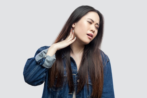 Dor de pescoço. Retrato de uma jovem asiática morena bonita doente em jaqueta jeans azul casual em pé sentindo desconforto e tocando seu pescoço. tiro de estúdio interior, isolado em fundo cinza claro.