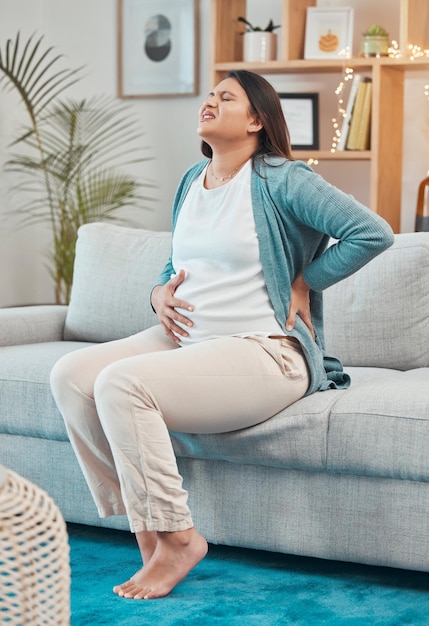 Dor nas costas grávida e mulher com dor e tensão corporal enquanto está sentada no sofá da sala Anatomia da barriga e desconforto na gravidez com uma mãe dolorosa infeliz com a condição física em casa