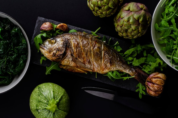 Dorada o pescado dorada al horno en un plato negro con rúcula y vegetales verdes sobre fondo negro