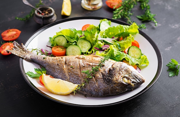 Dorado de pescado al horno con limón y ensalada fresca en plato blanco sobre fondo rústico oscuro. Cena saludable con concepto de pescado. Hacer dieta y comer limpio