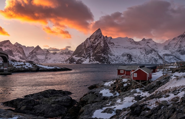 Dorf mit roten Hütten durch das Meer mit Bergen im Hintergrund bedeckt im Schnee im Sonnenuntergang in den Lofoten-Inseln, Norwegen