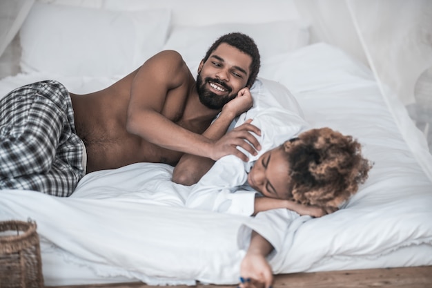 Dormilón. Sonriente joven afroamericano barbudo y esposa durmiendo profundamente en camisa blanca acostado en la cama en casa