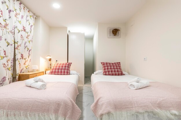Dormitorio con dos camas con mantas rosas, cojines rojos, toallas  enrolladas y llamativos visillos en un apartamento de alquiler vacacional