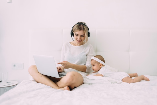 En el dormitorio, una joven y hermosa rubia está trabajando en una cama blanca con auriculares y una laptop junto a ella es una pequeña hija de 6 meses