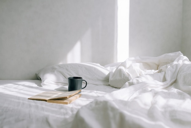 Dormitorio ligero y acogedor, taza de café o té y un libro abierto en la cama.