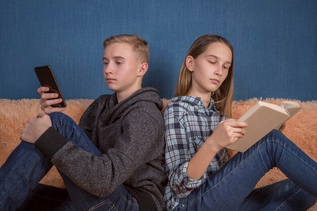 Foto dos adolescente y una niña leyendo juntos en interiores utilizando un teléfono y un libro de papel.