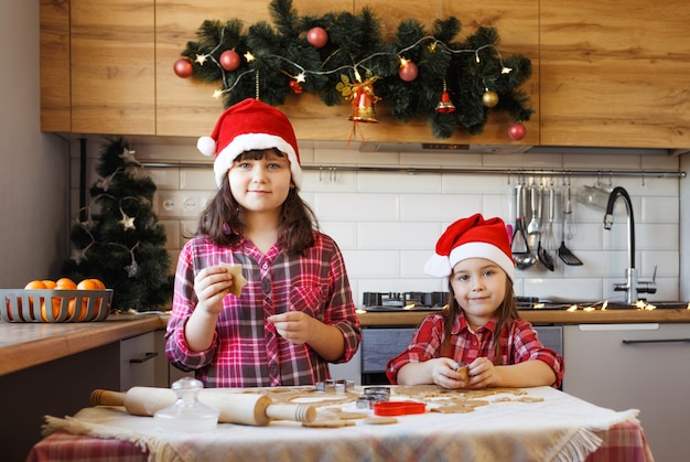 Dos adolescentes con camisas rojas a cuadros y sombreros navideños están haciendo galletas de jengibre con masa.