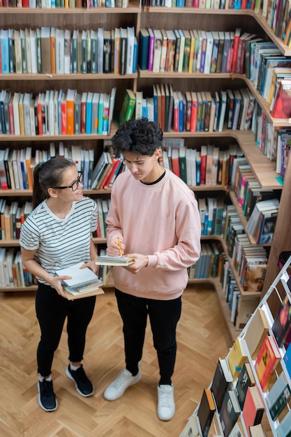 Dos adolescentes casuales de pie en la biblioteca de la universidad entre estantes con libros y discutiendo la tarea en casa después de clases