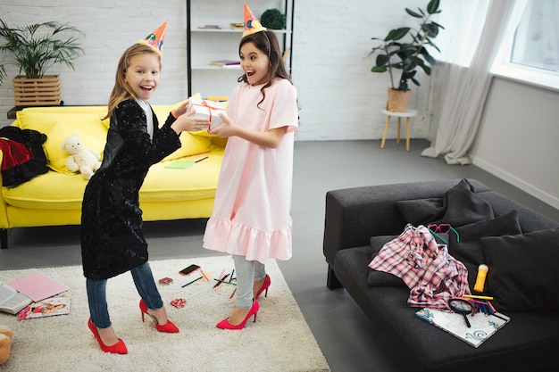 Dos adolescentes se divierten. Se paran en la habitación y sostienen un regalo juntos. Las niñas usan ropa y zapatos para mujeres adultas. Tienen fiesta de cumpleaños.