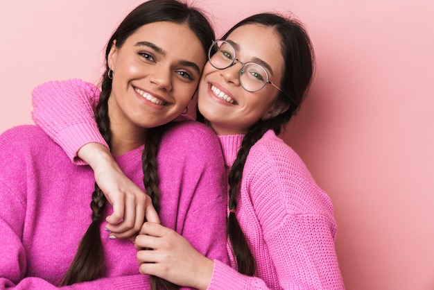 Dos adolescentes felices con trenzas en ropa casual sonriendo a la cámara aislada sobre pared rosa