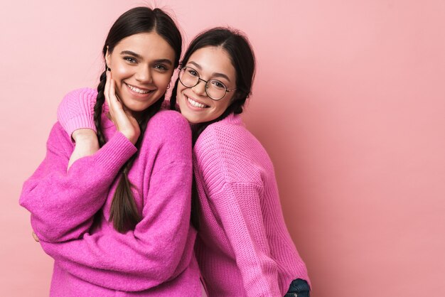 Dos adolescentes felices con trenzas en ropa casual sonriendo a la cámara aislada sobre pared rosa