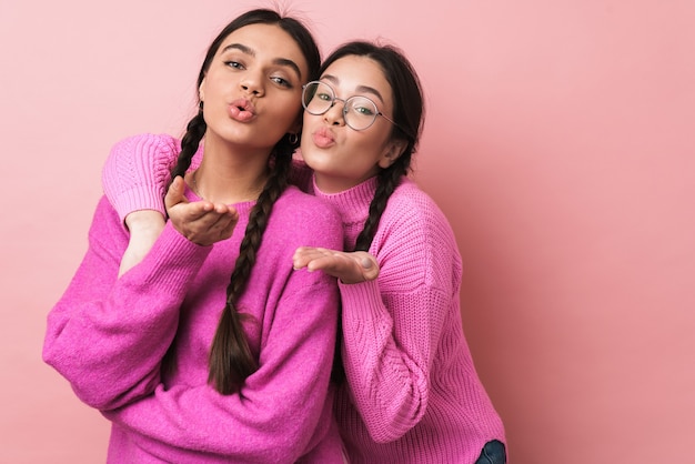 Dos adolescentes felices con trenzas en ropa casual sonriendo y soplando un beso de aire aislado sobre la pared rosa