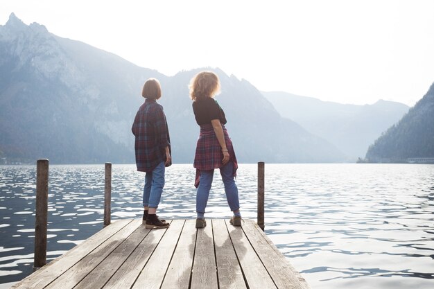 Dos adolescentes niñas sonrientes con soportes en un muelle en la orilla del lago y las montañas en el fondo