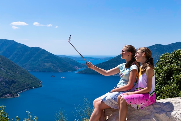Dos adolescentes en la toma de fotos por teléfono y palos de selfie en el fondo de la bahía y las montañas de Boko Kotorska en Montenegro