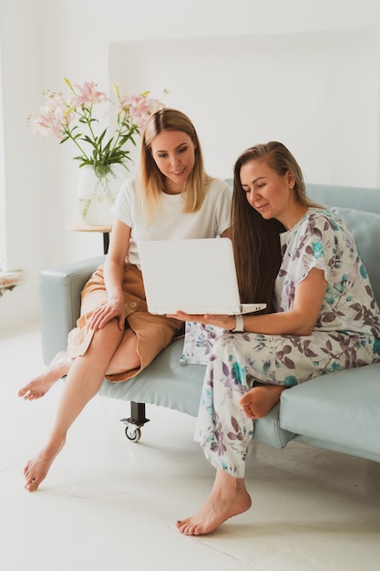 Dos adorables mujeres jóvenes charlando en casa en el sofá, tomando café y trabajando en una computadora portátil