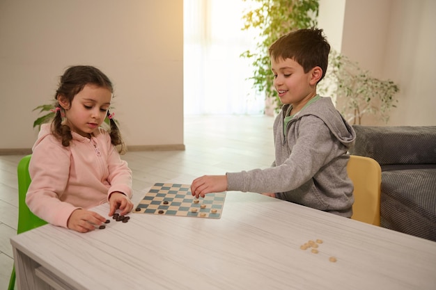 Dos adorables niños caucásicos de primaria, niño y niña, hermano y hermana que se divierten jugando al juego de tablero de damas juntos en el interior de la casa.