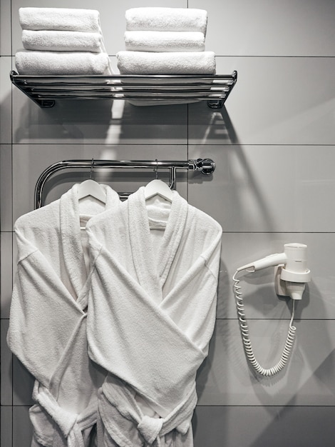 Dos albornoces blancos en el baño en la pared con un secador de pelo blanco y toallas en el hotel.