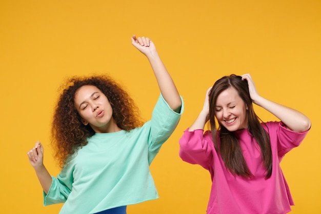 Dos alegres amigas europeas afroamericanas vestidas de verde rosa posando aisladas en un fondo amarillo. Concepto de estilo de vida de las personas. Simulacros de espacio de copia. Divertirse bailando levantando las manos.