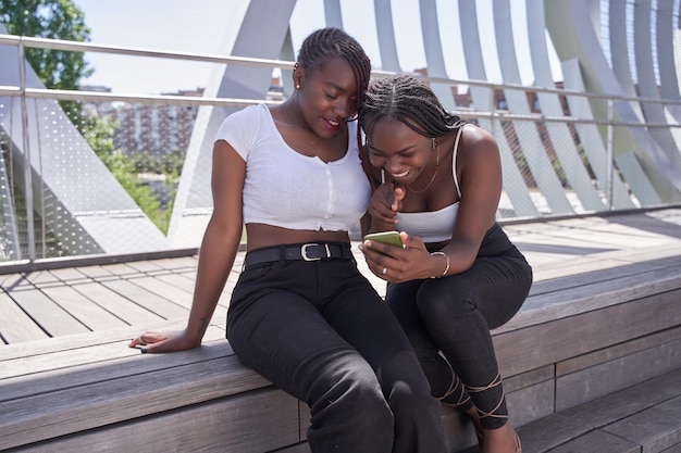 Dos alegres mujeres afroamericanas están sentadas juntas viendo algo por teléfono