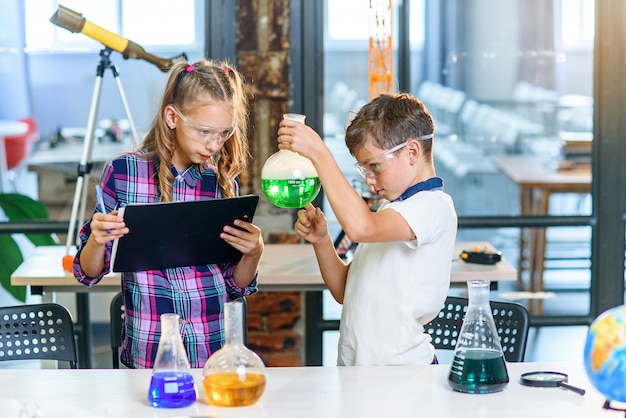 Dos alumnos de primaria en gafas protectoras haciendo experimentos con líquidos coloreados en frascos de vidrio y hielo seco.