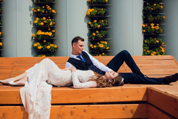 Dos amantes se sientan en un banco, los recién casados se agacharon para descansar en los brazos del otro durante una sesión de fotos de boda, la novia con un vestido blanco y el novio con un hermoso traje retirado en el parque.