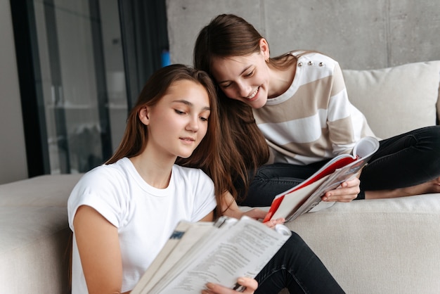 Dos amigas adolescentes alegres sentados en casa en un sofá leyendo revistas