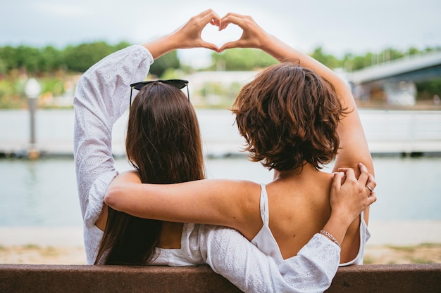 Dos amigas adolescentes haciendo forma de corazón con sus manos mientras están sentadas en un banco junto a un río