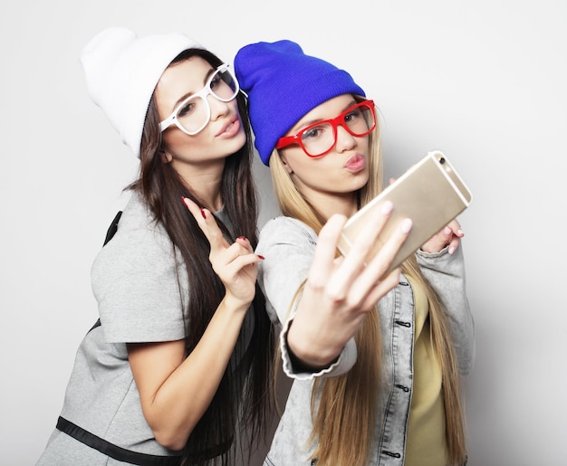 Dos amigas adolescentes en traje hipster hacen selfie sobre fondo blanco.