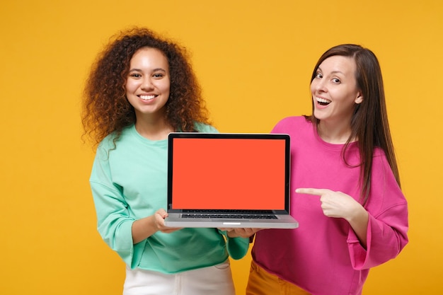 Dos amigas africanas europeas con ropa verde rosa posando aisladas en un fondo amarillo. Concepto de estilo de vida de las personas. Simulacros de espacio de copia. Señale con el dedo en la computadora portátil con pantalla vacía en blanco.