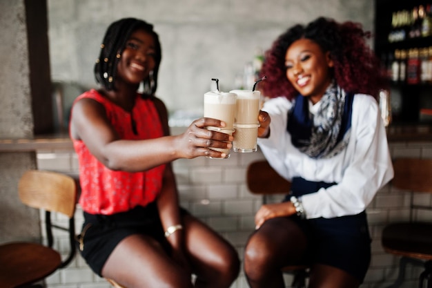 Dos amigas afroamericanas atractivas sentadas en sillas contra el bar en el pub y bebiendo café con leche