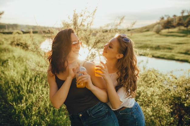 Dos amigas agradables están tomando un cóctel con paja, divirtiéndose al atardecer, expresión facial positiva, al aire libre