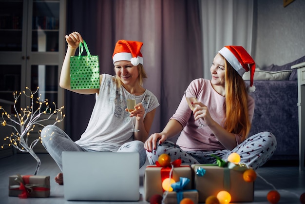 Dos amigas alegres abren cajas de regalo de Navidad durante la videoconferencia en línea con la familia en la computadora portátil. Las hermanas intercambian regalos en el interior de la casa con decoraciones festivas.