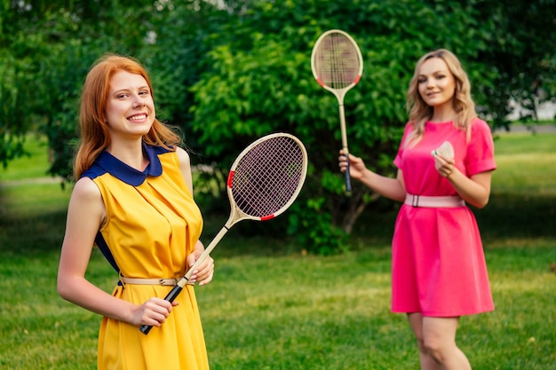 Dos amigas alegres divertidas hermosa joven pelirroja pelirroja irlandesa con un vestido amarillo y una persona femenina rubia europea con un vestido rosa jugando raqueta de bádminton de tenis en el parque de verano