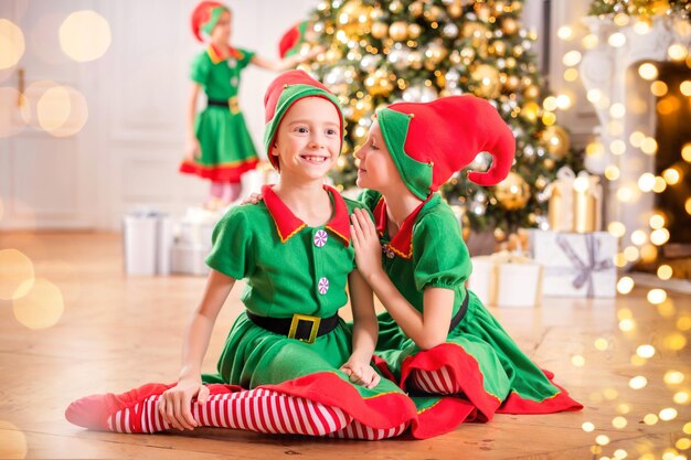 Dos amigas alegres y felices disfrazadas de duendes de Papá Noel en el fondo de un árbol de Navidad y otros niños