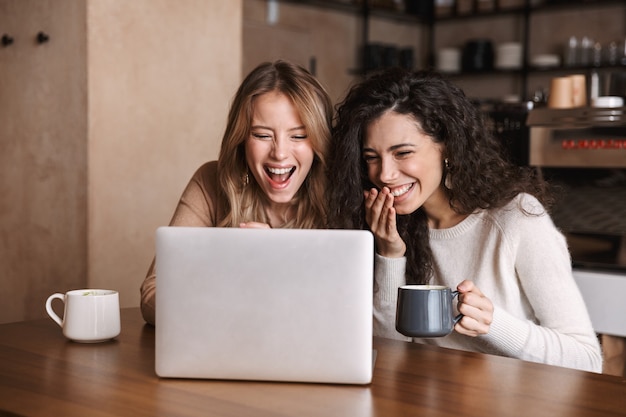 Dos amigas alegres jóvenes sentados en la mesa de café, divirtiéndose juntos, tomando café, usando la computadora portátil