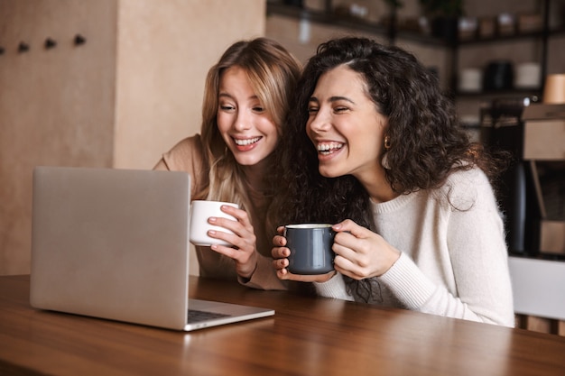 Dos amigas alegres jóvenes sentados en la mesa de café, divirtiéndose juntos, tomando café, usando la computadora portátil