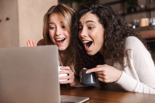 Dos amigas alegres jóvenes sentados en la mesa de café, divirtiéndose juntos, tomando café, usando la computadora portátil