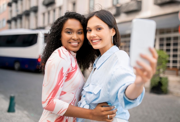 Dos amigas alegres con teléfonos inteligentes tomando selfies mientras caminan al aire libre