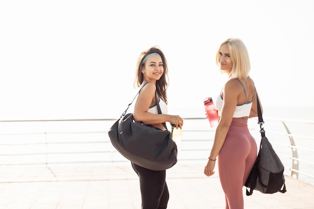Dos amigas atléticas en ropa deportiva con esteras de yoga bolsas y botella de agua en la playa estilo de vida saludable