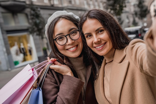 Dos amigas con bolsas de compras haciendo selfie afuera en la ciudad