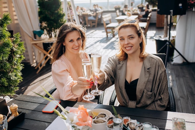 Dos amigas comiendo ostras frescas y bebiendo vino prosecco frío en el atardecer de verano en el restaurante Delicias de mariscos