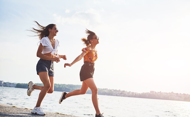 Dos amigas corren y se divierten en la playa cerca del lago durante el día soleado.