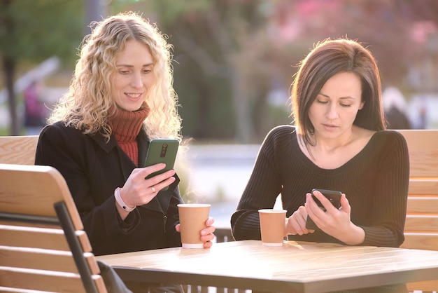 Dos amigas distraídas navegando por sus teléfonos móviles mientras se sientan juntas en un café de la calle de la ciudad Novias ignorándose mientras miran dispositivos Adicción del concepto de redes sociales