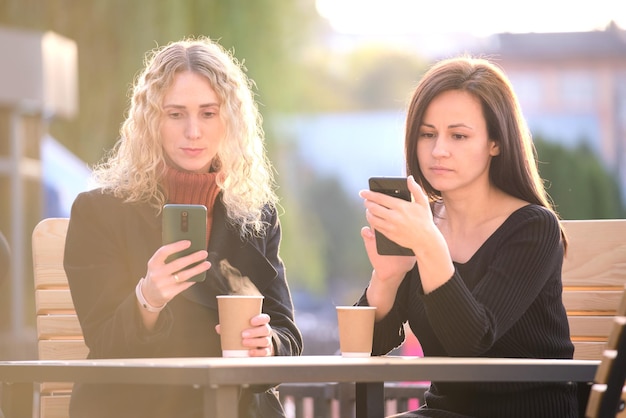 Dos amigas distraídas navegando por sus teléfonos móviles mientras se sientan juntas en un café de la calle de la ciudad Novias ignorándose mientras miran dispositivos Adicción del concepto de redes sociales