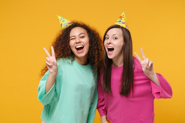 Dos amigas divertidas chicas afroamericanas europeas con ropa verde rosa, sombreros de cumpleaños posando aisladas en un fondo amarillo. Concepto de estilo de vida de las personas. Simulacros de espacio de copia. Mostrando el signo de la victoria.