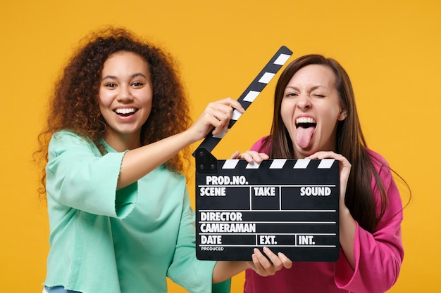 Dos amigas divertidas europeas afroamericanas vestidas de verde rosa posando aisladas en un fondo amarillo. Concepto de estilo de vida de las personas. Simulacros de espacio de copia. Sujeta la clásica claqueta negra para hacer películas.