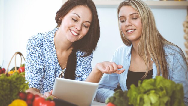 Dos amigas eligiendo la receta de una deliciosa comida mientras están sentadas en la mesa de la cocina Tablet pc es el mejor libro de cocina